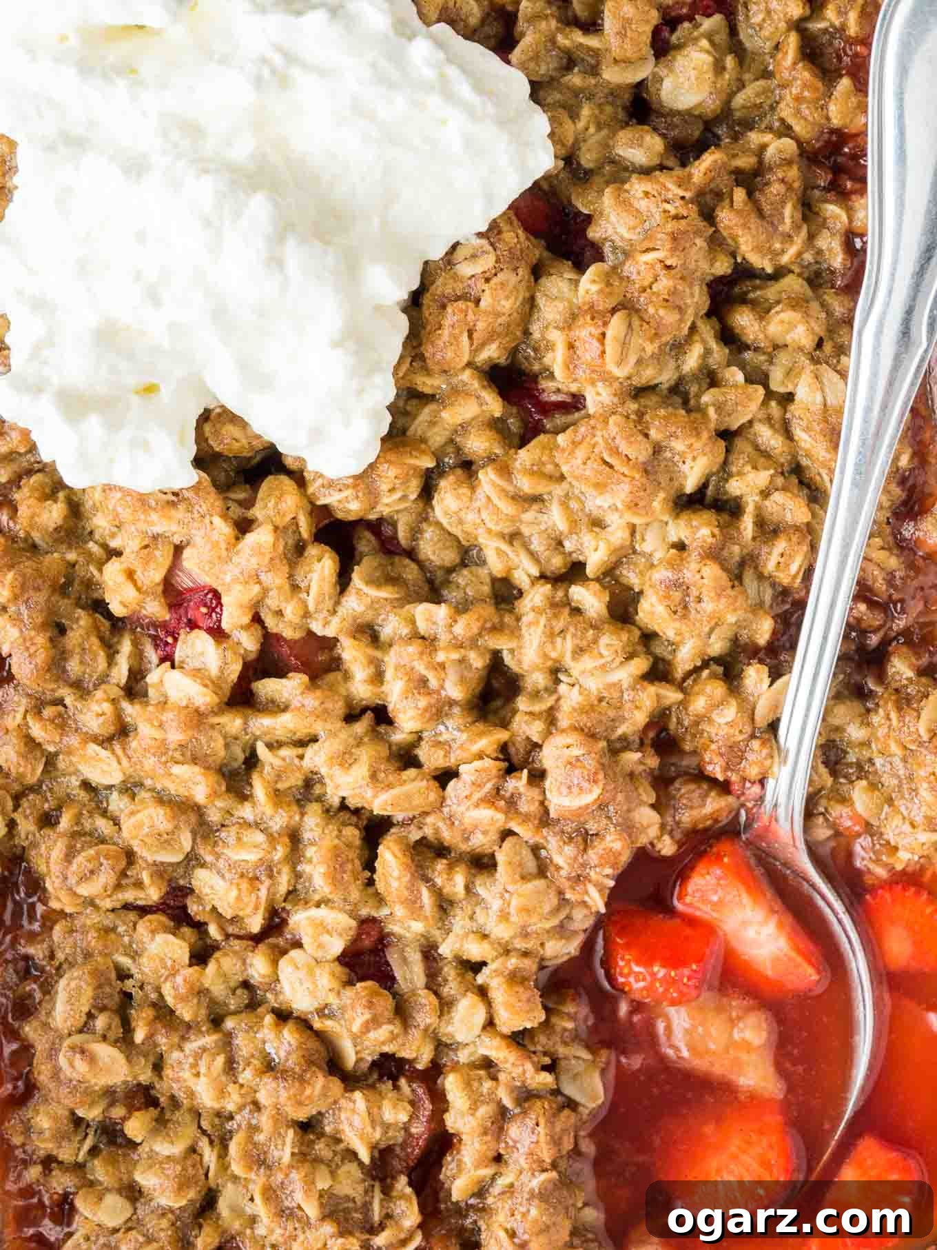 A close-up shot of Strawberry Rhubarb Crisp, showing a spoon digging into the warm, bubbly fruit layer beneath the golden-brown, crunchy topping, garnished with fresh cream.