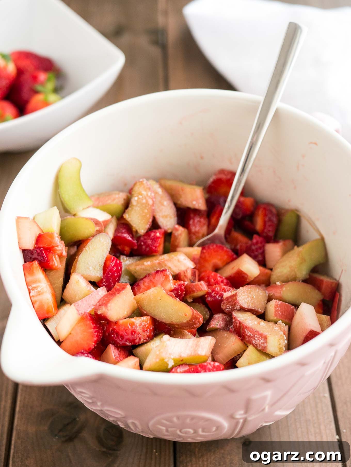 Freshly cut strawberries and rhubarb pieces artfully arranged in a pink bowl with a fork, with whole fresh strawberries in a rustic bowl in the background.