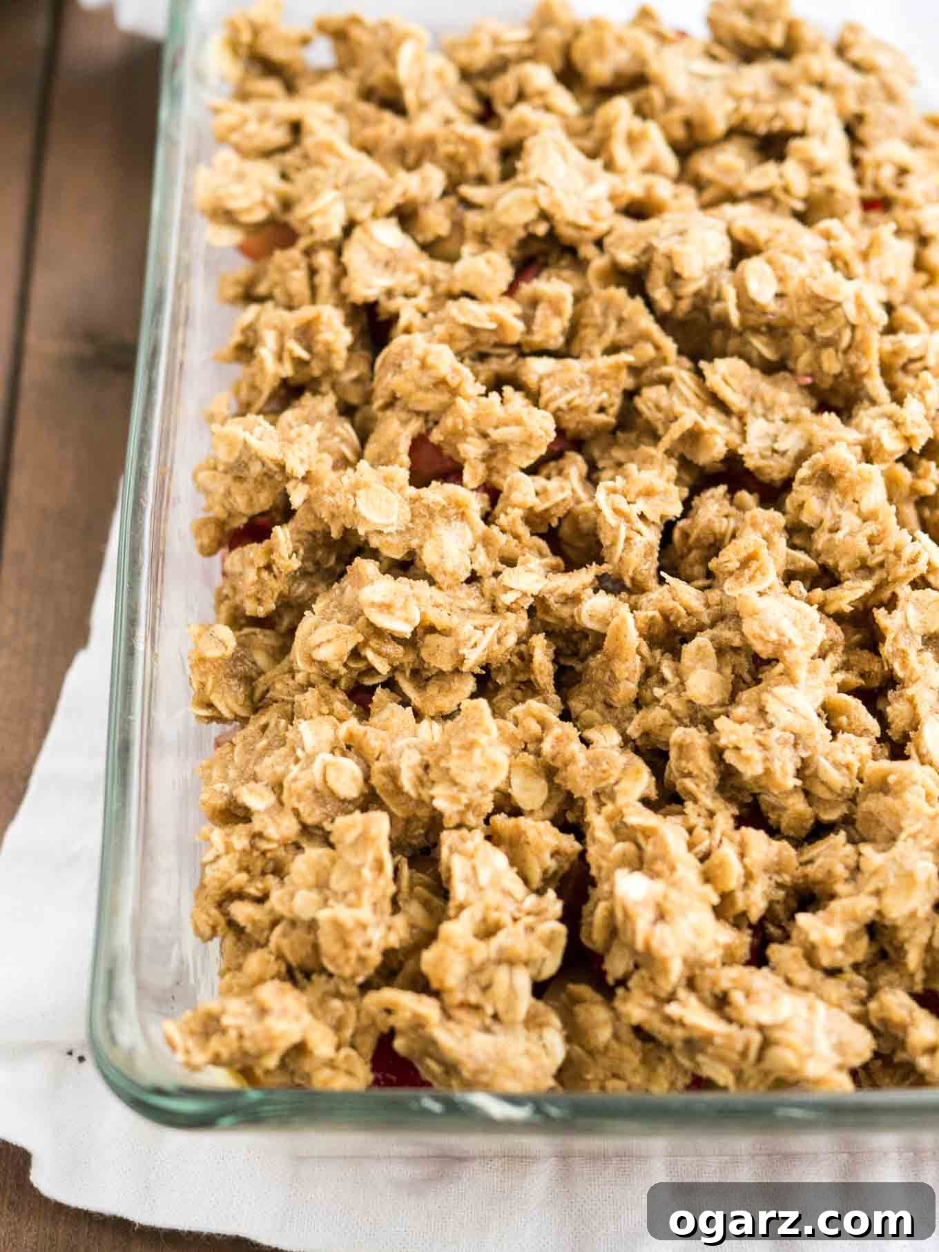 A glass baking dish filled with Strawberry Rhubarb Crisp, showcasing the unbaked oatmeal streusel topping covering the fruit layer, placed on a rustic wooden table.