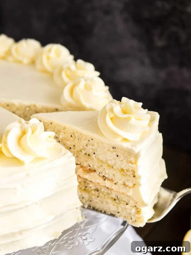 Close-up of a glass serving platter with a lemon poppy seed cake on a wooden table with a white tablecloth.