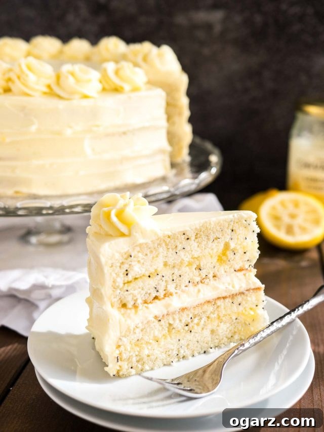 A slice of lemon poppy seed cake sitting on a white plate with a fork. The rest of the cake is on a glass serving platter in the background next to some lemons.