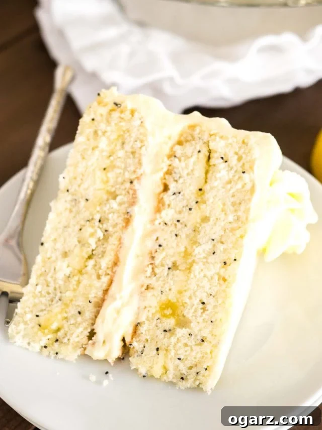 A slice of lemon poppy seed cake lyingon a white plate with a fork. Tere\'s a white dishcloth and a lemon next to it. 