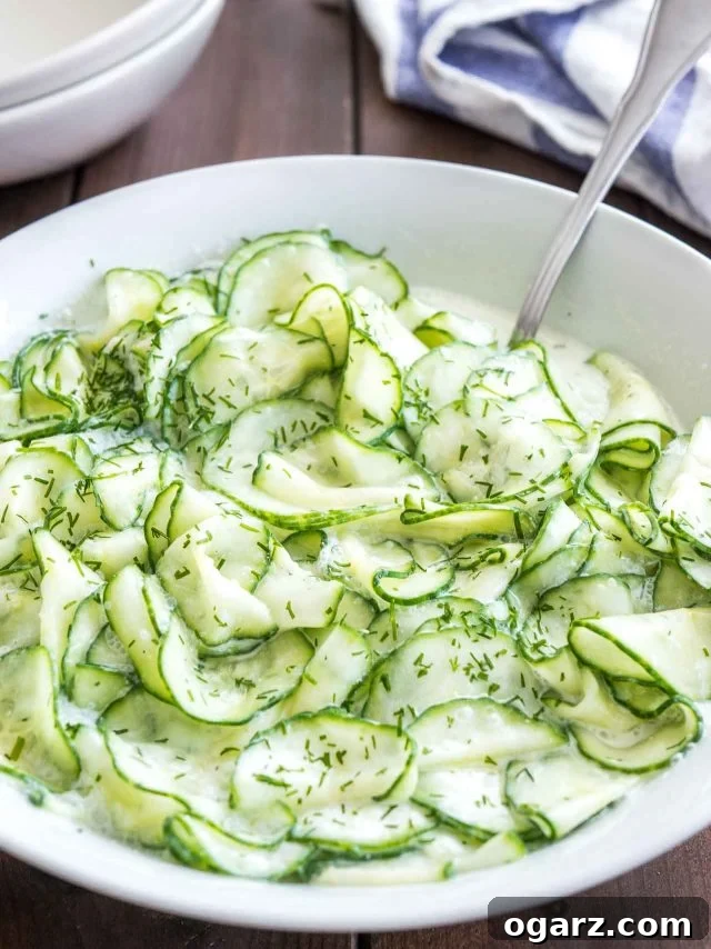 Refreshing German Cucumber Salad 2 A pristine white salad bowl filled with German Cucumber Salad, garnished with fresh dill, sits on a rustic wooden table. A spoon rests in the bowl, inviting a taste of this creamy summer delight.