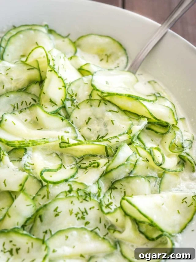 Refreshing German Cucumber Salad 3 A close-up view of German Cucumber Salad in a white bowl, showcasing the thinly sliced cucumbers coated in a creamy dill dressing. A spoon is ready for serving.