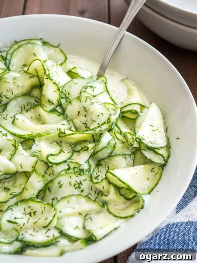 Refreshing German Cucumber Salad 4 A close-up of German Cucumber Salad in a white bowl, resting on a blue and white patterned dish towel. A spoon is positioned for serving, highlighting the salad's fresh and inviting texture.