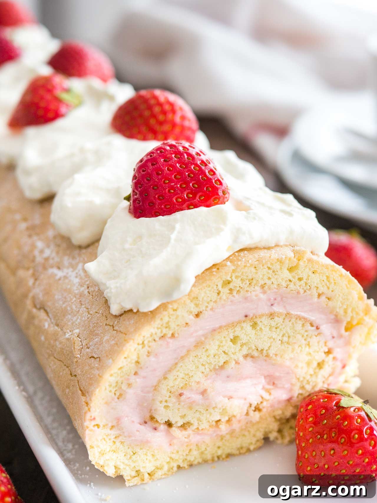 Strawberry Dream Roll 2 Close-up of a beautifully sliced strawberry cake roll, adorned with fresh whipped cream and vibrant strawberries, presented on an elegant rectangular serving platter. The cake reveals its delightful swirl of sponge and creamy filling.