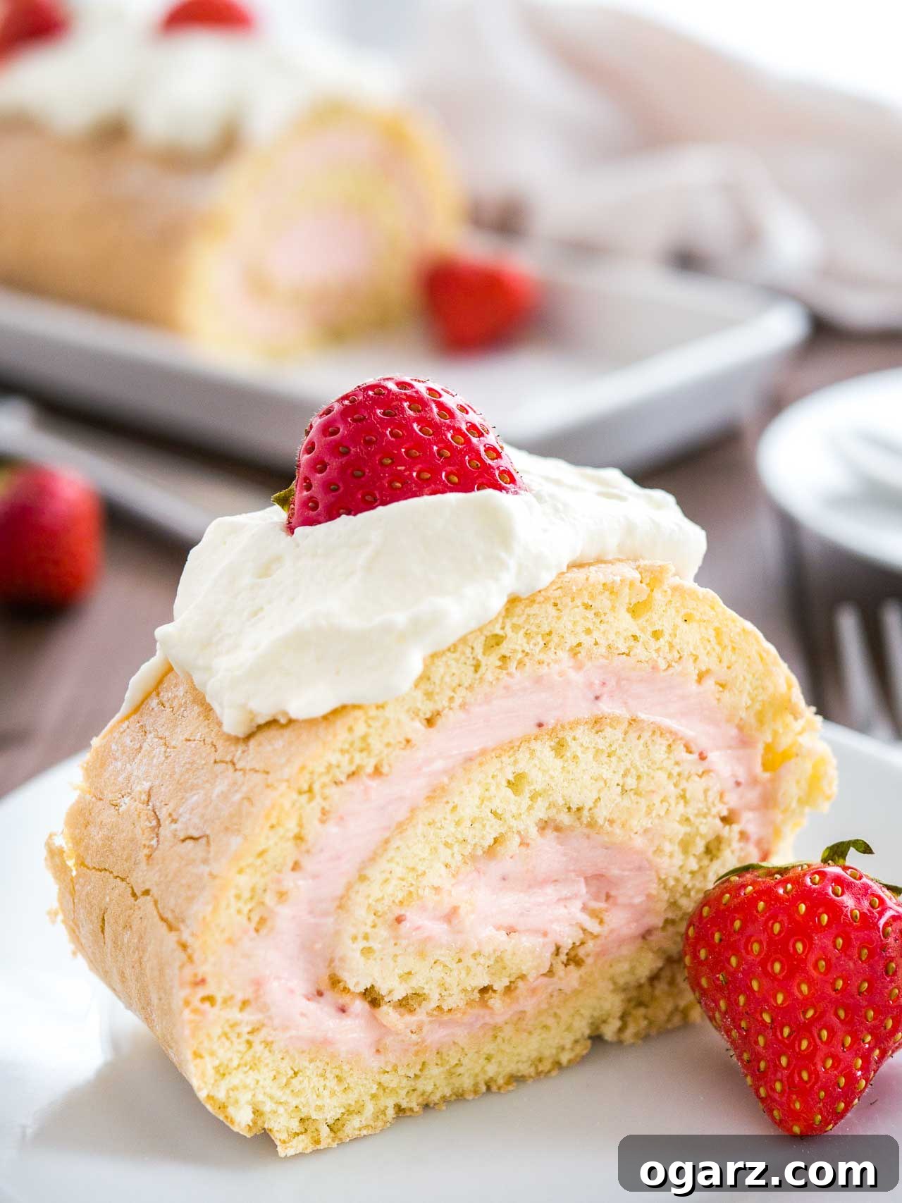 Strawberry Dream Roll 6 A perfectly portioned slice of strawberry swiss roll cake, artfully arranged on a pristine white plate and crowned with a dollop of whipped cream and a fresh strawberry. The remainder of the elegant roll cake rests invitingly in the background.