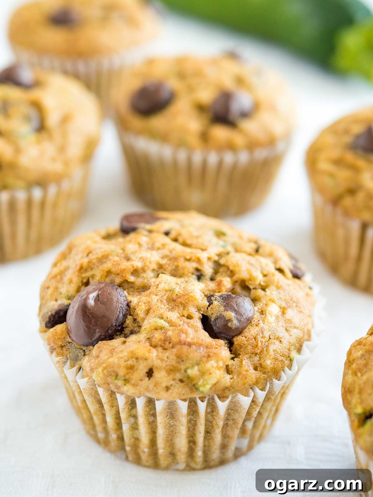 Chocolate Zucchini Delight Muffins 3 Close-up of a stack of zucchini chocolate chip muffins on a white tablecloth, with blurred fresh zucchini in the background, highlighting the texture and chocolate chips.
