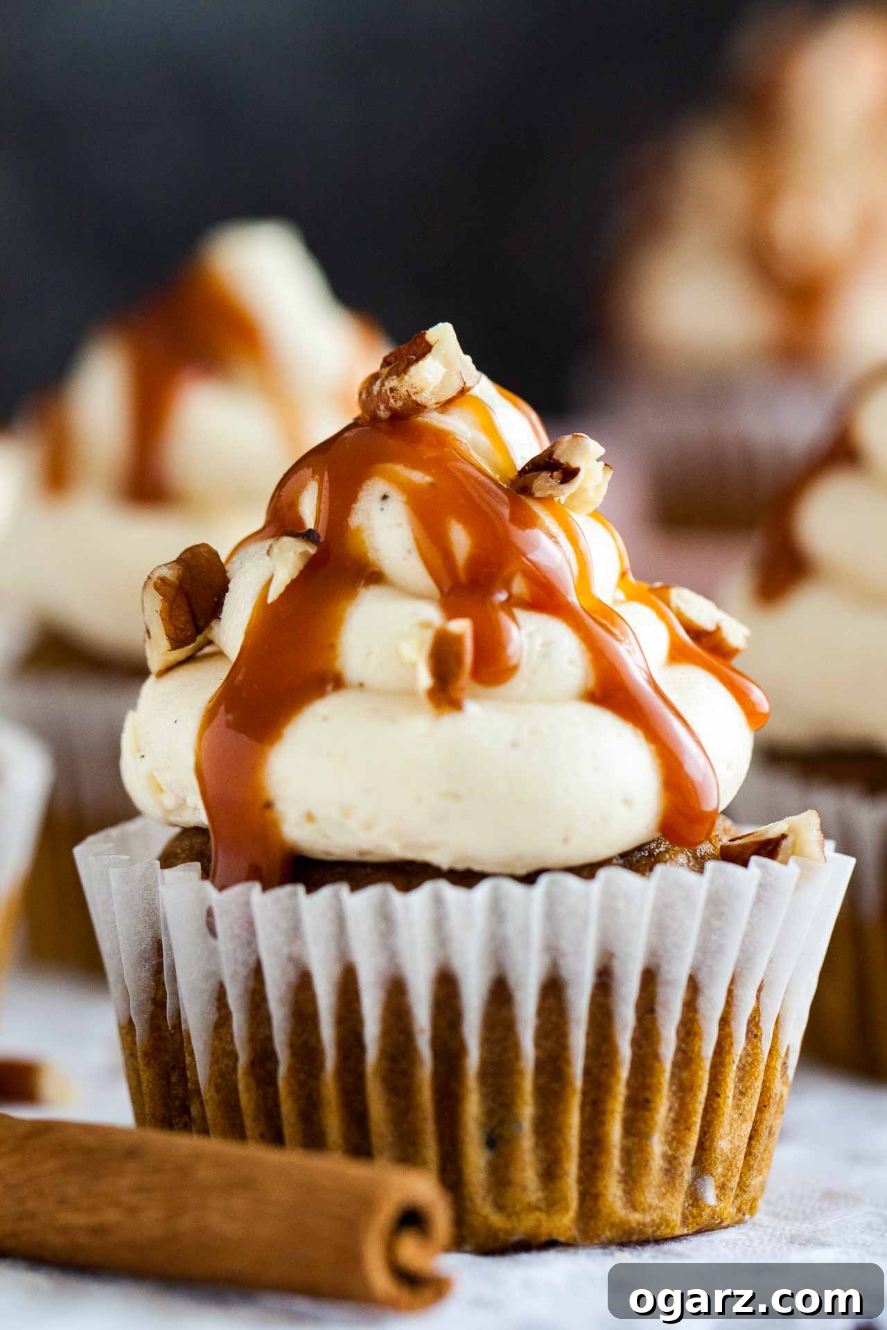 Delicious pumpkin cupcake with brown butter frosting, salted caramel drizzle, and pecan nuts, resting on a rustic grey tablecloth next to a cinnamon stick.