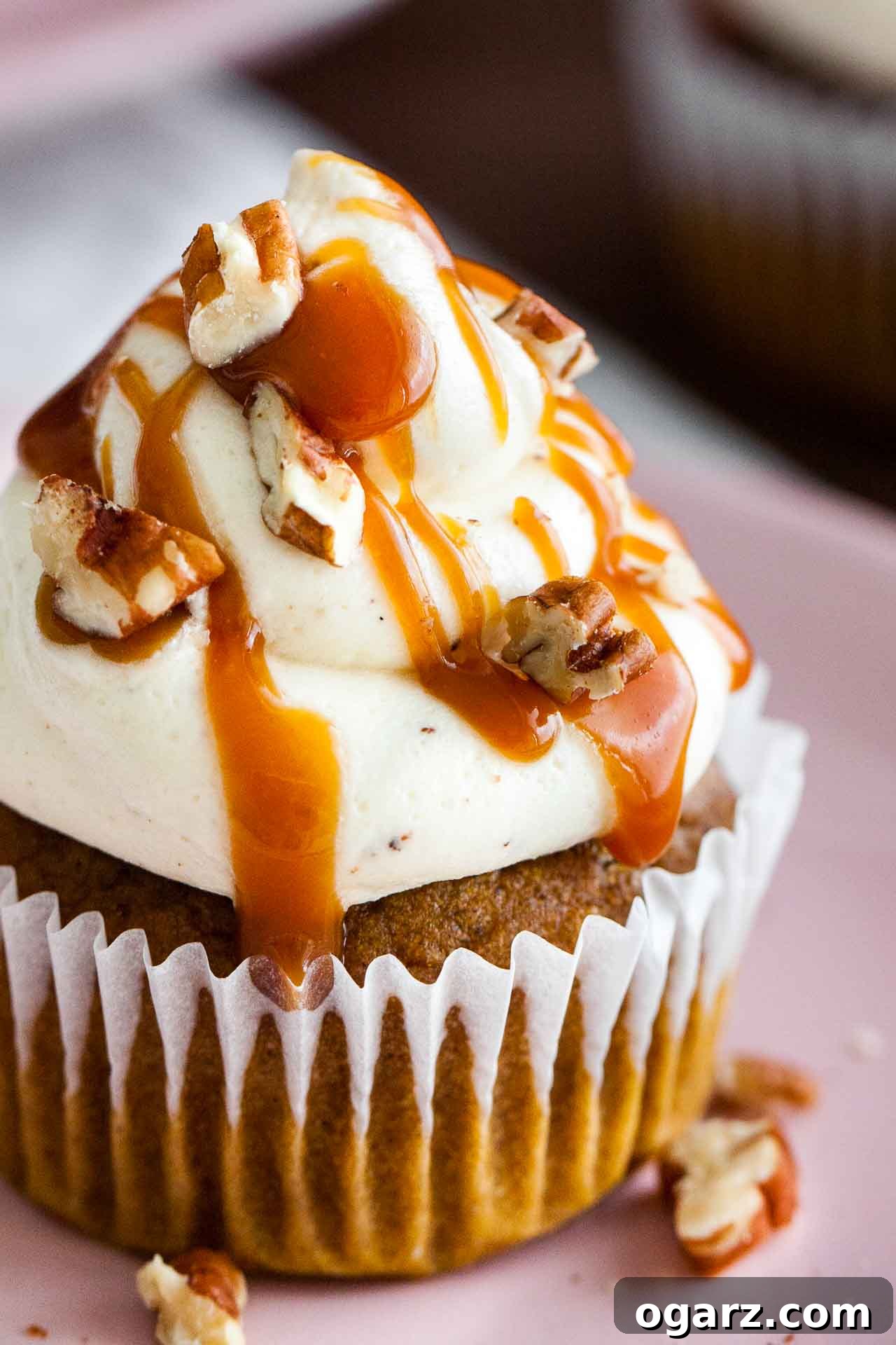 A pair of perfectly baked pumpkin cupcakes, each crowned with fluffy brown butter frosting, glistening caramel sauce, and crunchy pecan pieces, served on a vibrant teal plate.