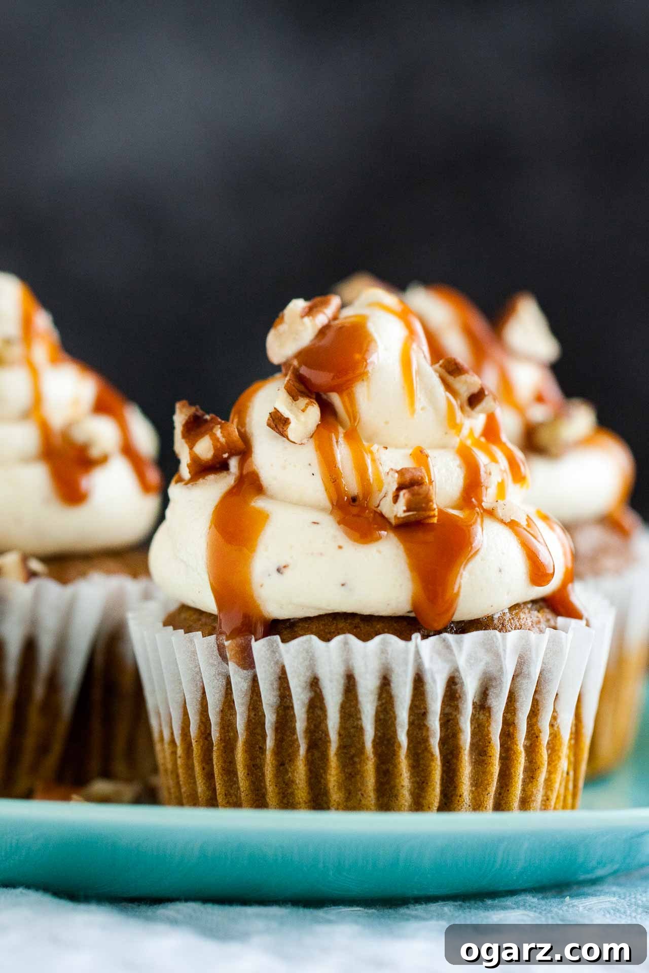 One exquisitely frosted pumpkin cupcake with brown butter and caramel on a pink plate, with more autumnal cupcakes softly blurred in the background.