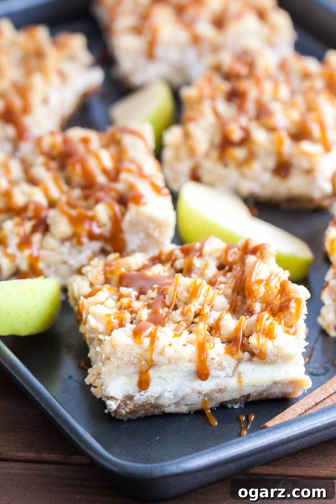 Salted Caramel Apple Cheesecake Bars 3 A black baking sheet with caramel apple cheesecake bars, garnished with apple wedges and cinnamon sticks.
