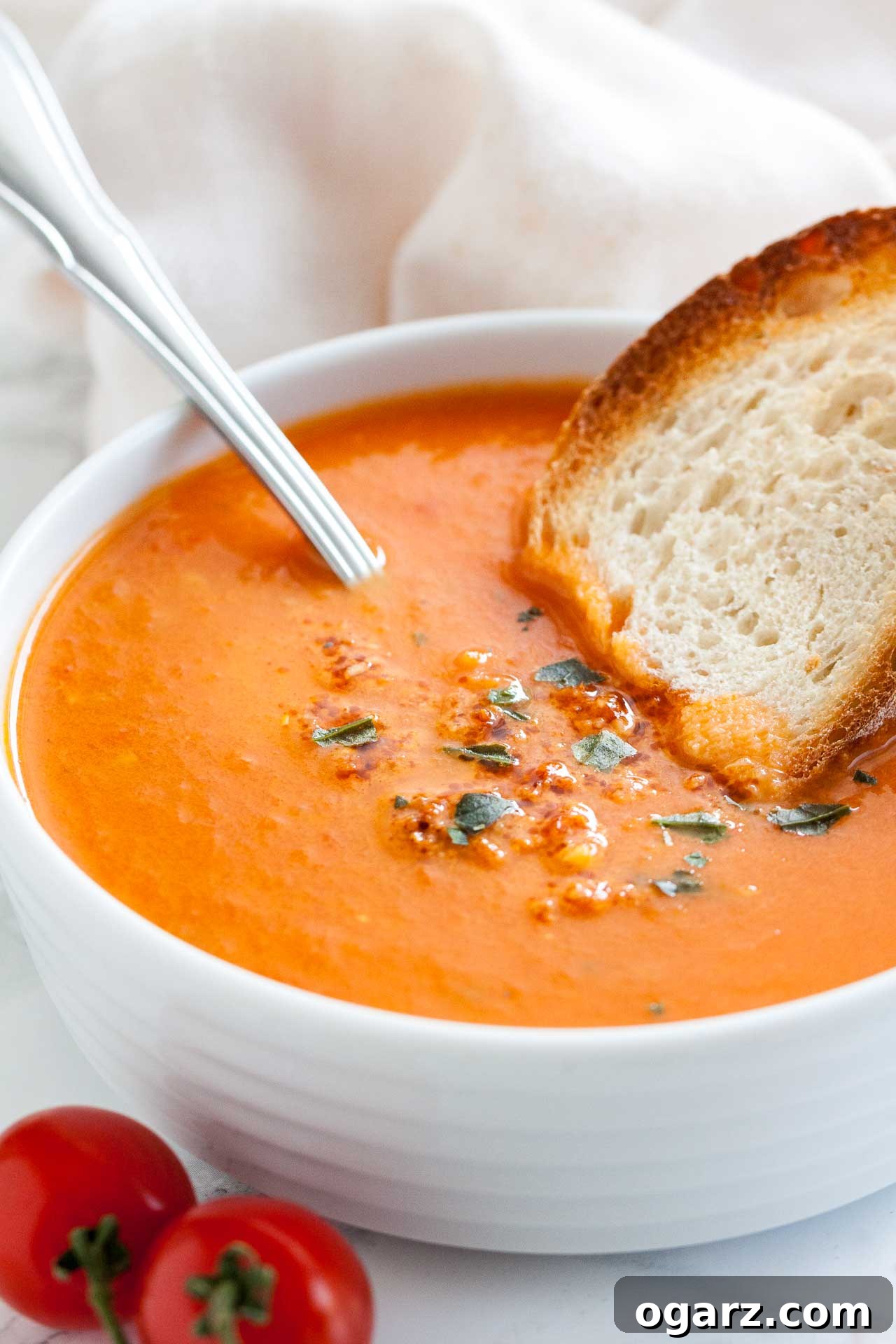 Golden Roasted Tomato Velouté 2 A white bowl of roasted tomato soup garnished with basil, with a spoon and a toasted slice of bread on a white dishtowel.