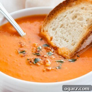 Golden Roasted Tomato Velouté 6 A white bowl of roasted tomato soup garnished with basil, with a spoon and a toasted slice of bread on a white dishtowel.