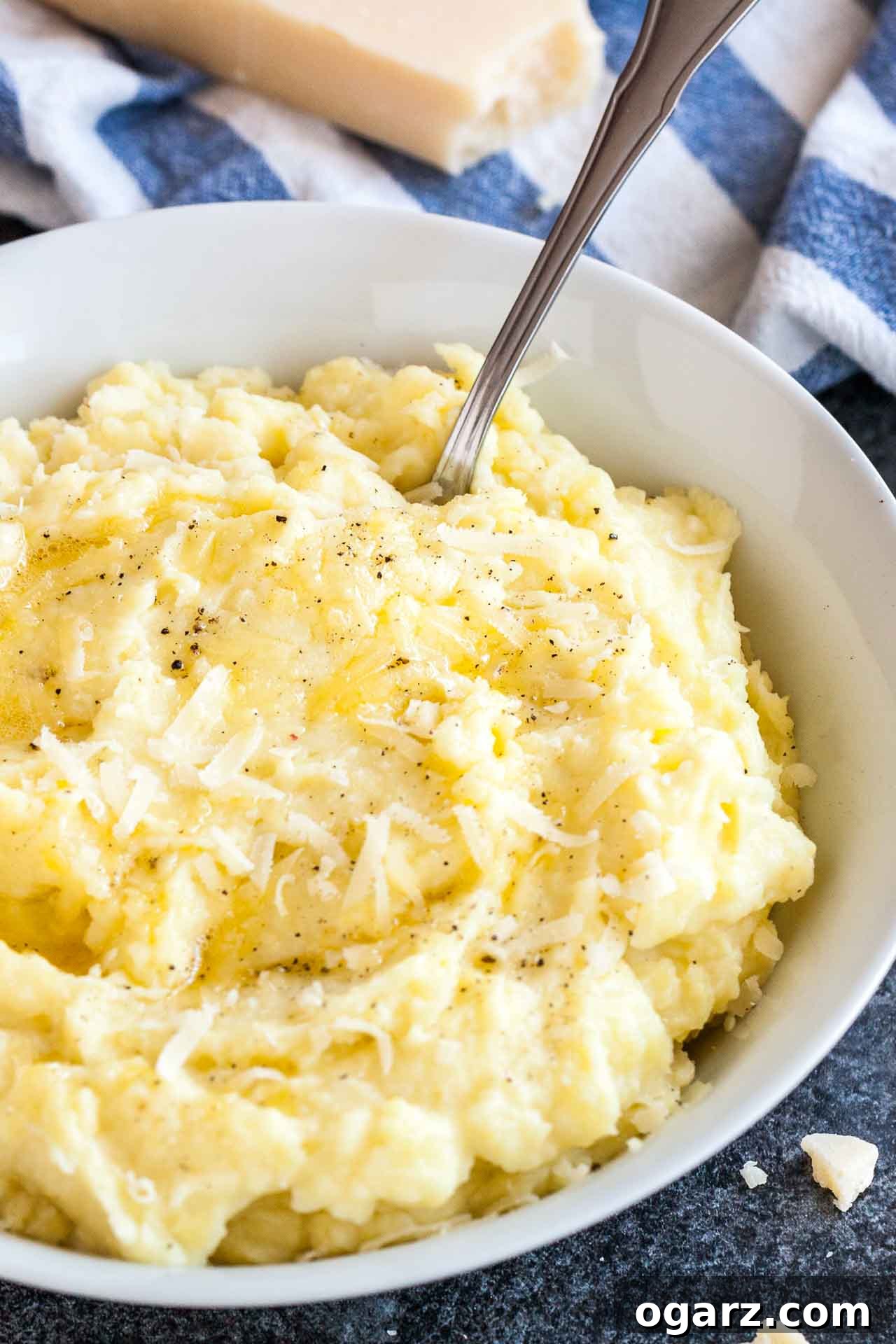 Golden Garlic Mash 2 A beautifully presented white bowl of roasted garlic mashed potatoes, generously topped with freshly grated Parmigiano cheese and a sprinkle of black pepper. A spoon is gently resting in the creamy potatoes, with a crisp white and blue dishtowel and a block of Parmigiano in the background, hinting at fresh ingredients.
