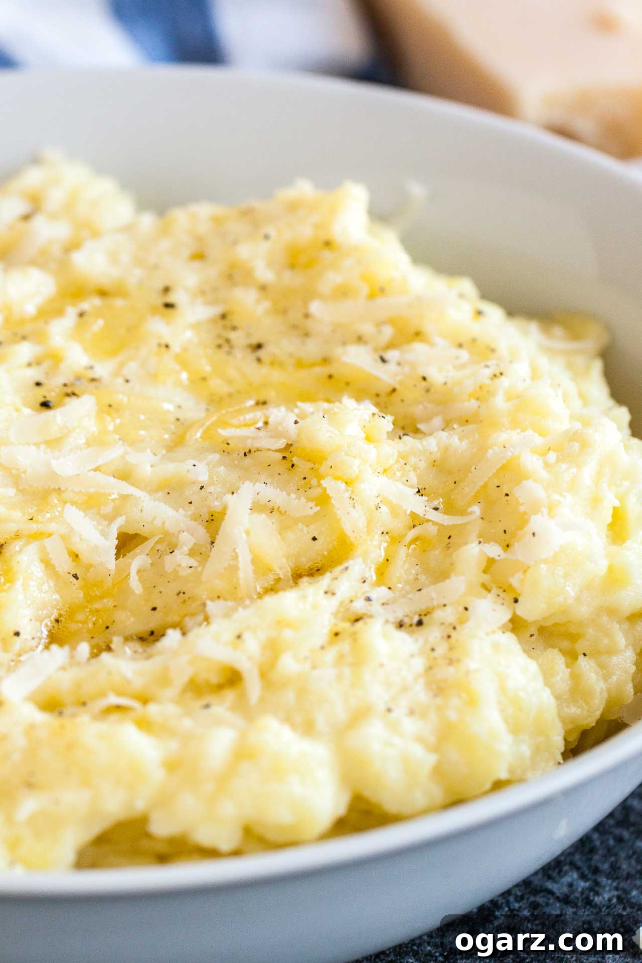 Golden Garlic Mash 3 A pristine white bowl filled with creamy roasted garlic mashed potatoes, artfully garnished with a generous sprinkle of Parmigiano cheese and a dusting of ground black pepper. In the soft-focus background, a white and blue patterned dishtowel adds a touch of rustic charm, alongside a solid block of Parmigiano cheese, emphasizing freshness and quality.