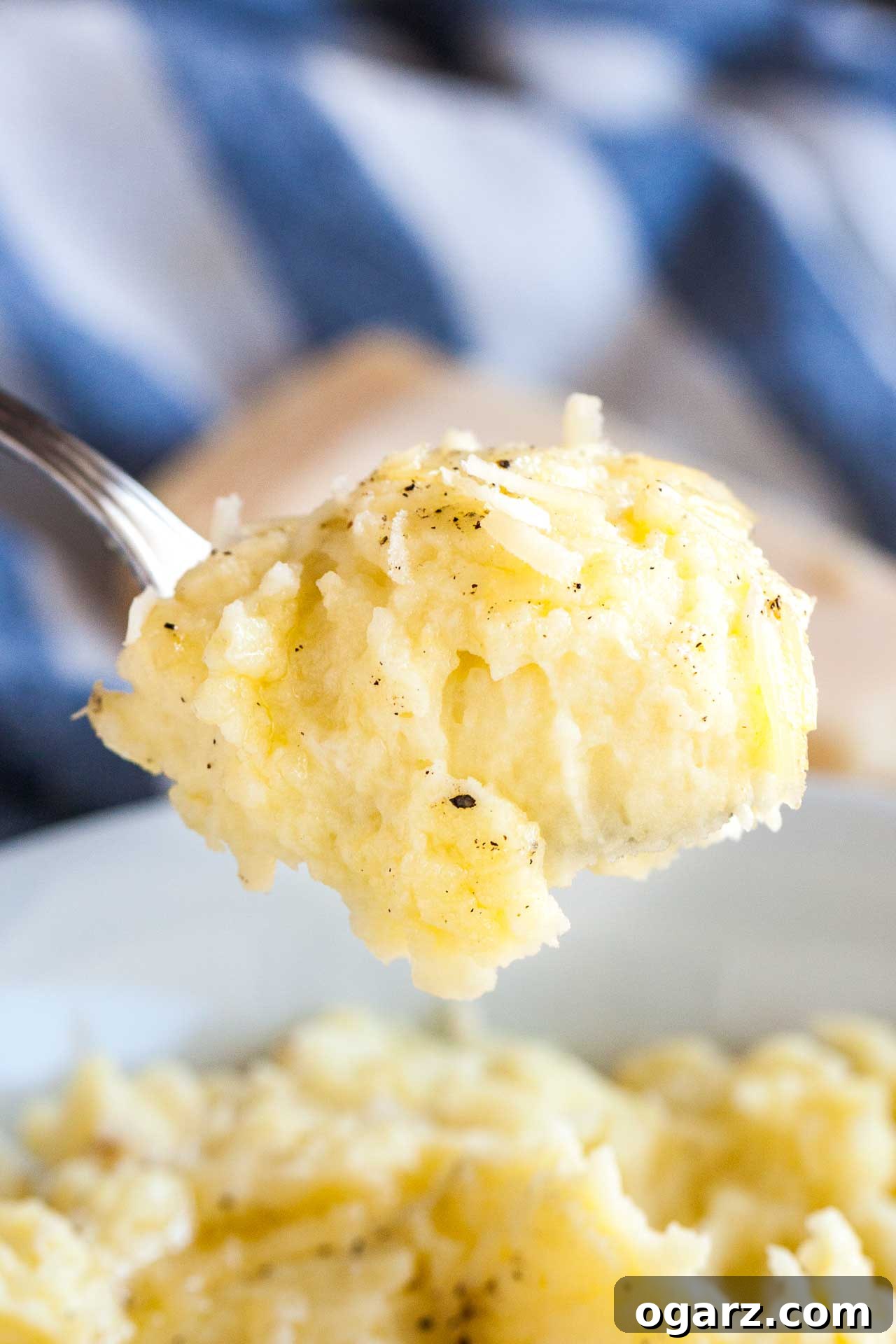 Golden Garlic Mash 4 A close-up shot of a spoon expertly holding up a luscious dollop of roasted garlic mashed potatoes, suspended above a white bowl filled with more creamy mashed potatoes. In the soft background, a white and blue dish towel and a block of Parmigiano cheese complete the inviting scene, suggesting fresh ingredients and homemade goodness.