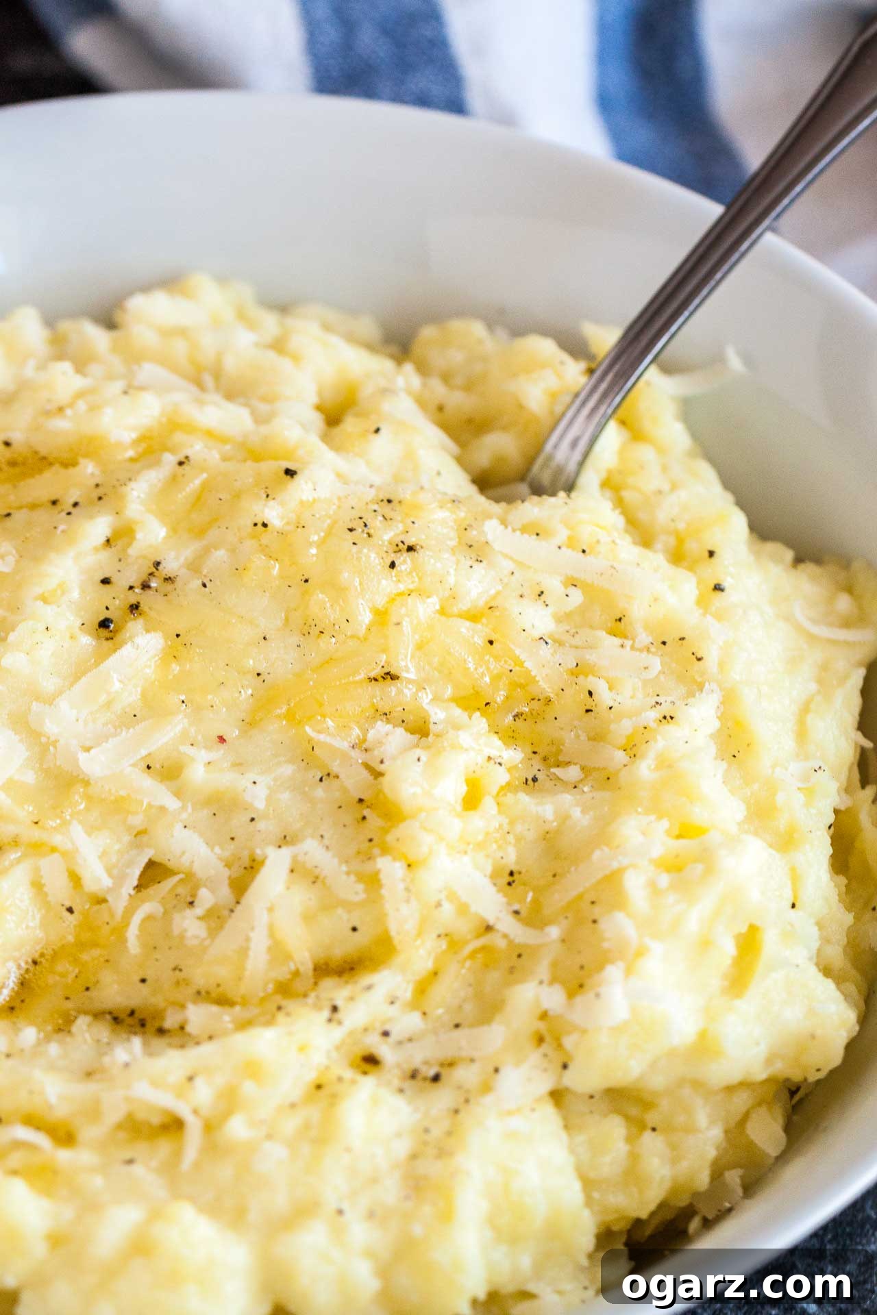 Golden Garlic Mash 5 A close-up and inviting view of a white bowl filled with incredibly creamy roasted garlic mashed potatoes. The surface is beautifully topped with a generous sprinkle of freshly grated Parmigiano cheese and a dash of ground black pepper. A spoon is gently tucked into the potatoes, ready for tasting. In the blurred background, a white and blue dishtowel provides a cozy, homemade ambiance.