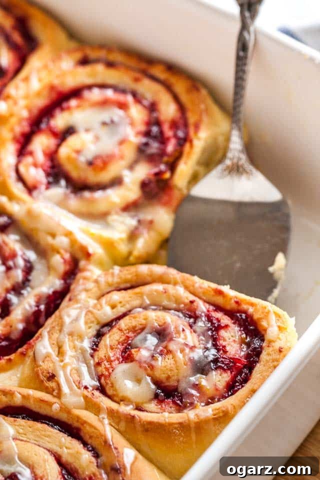 Zesty Cranberry Orange Buns 5 Close-up of a white baking dish with orange cranberry rolls. One of the rolls is missing and a cake server is lifting out another.