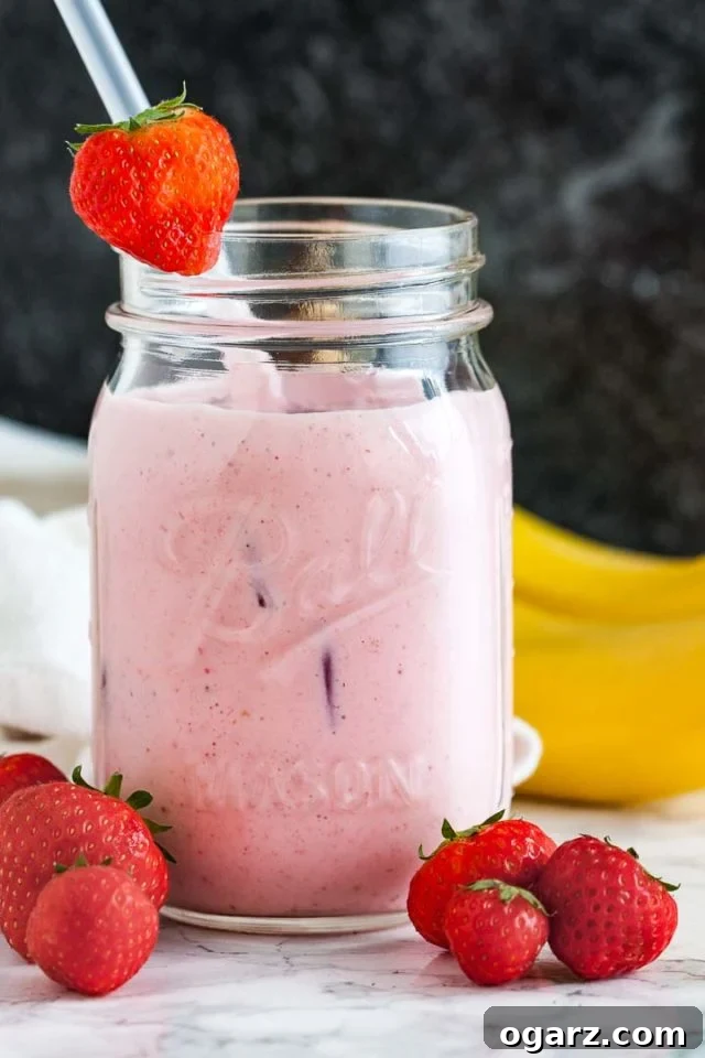 A ball jar with a strawberry banana smoothie a white straw and a strawberry on the rim on a marble surface in front of a dark background. There are some strawberries in front and 2 bananas in the back.