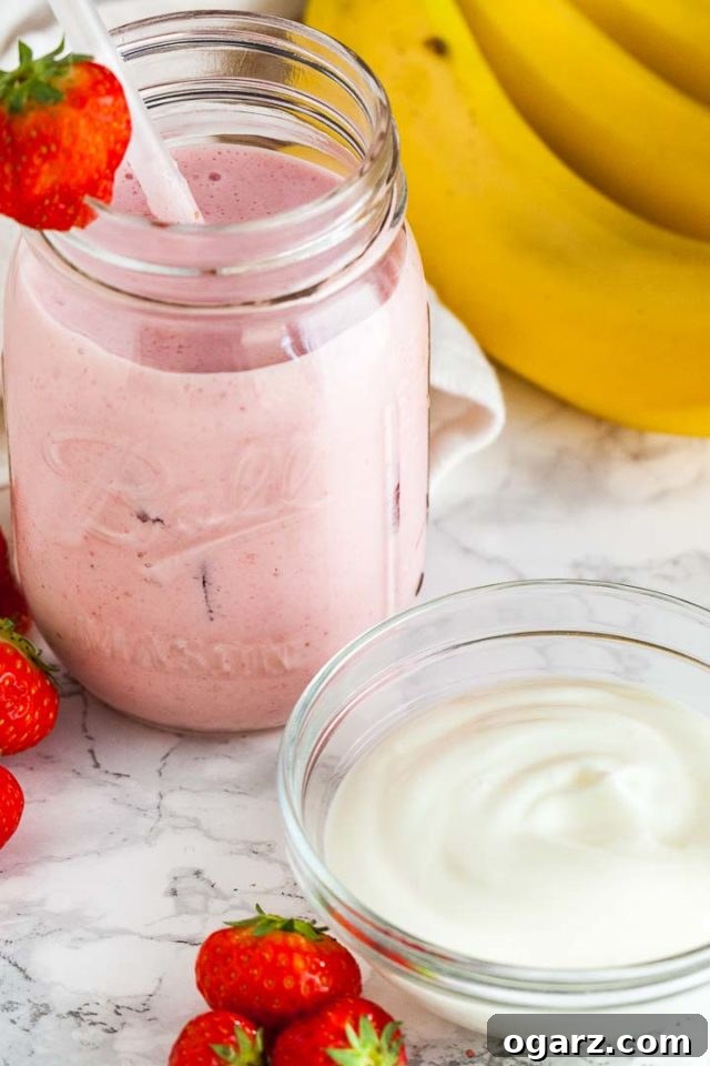 A ball jar with a strawberry banana smoothie a white straw and a strawberry on the rim on a marble surface. There are bananas, strawberries and a small glass bowl of yogurt next to it.