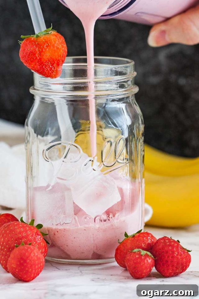 Strawberry banana smoothie being poured into a ball jar with ice, a straw and a strawberry on the rim in front of a dark background. There are some strawberries and 2 bananas next to it.