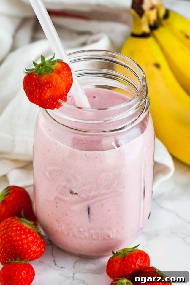 A ball jar with a strawberry banana smoothie a white straw and a strawberry on the rim on a marble surface. There's a white dish towel, 2 strawberries and some bananas next to it.