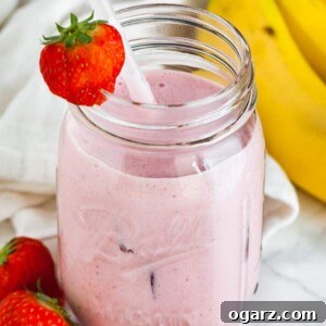 A ball jar with a strawberry banana smoothie a white straw and a strawberry on the rim on a marble surface. There's a white dish towel, 2 strawberries and some bananas next to it.