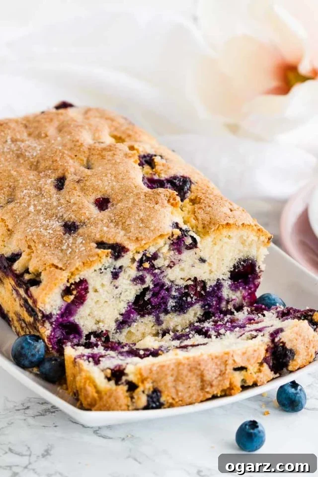 A loaf of blueberry bread on a white, rectangular plate with 1 slice cut off lying in front of it, garnished with blueberries and a flower.