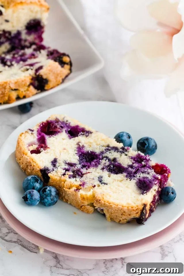 A slice of blueberry bread on a white plate stacked on a pink plate, with blueberries next to a rectangular plate with a slice of blueberry bread on it.