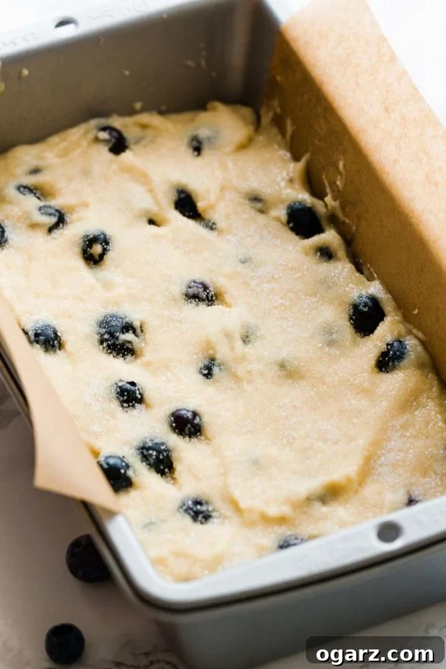 A cake pan, lined with parchment paper, filled with dough with blueberries.