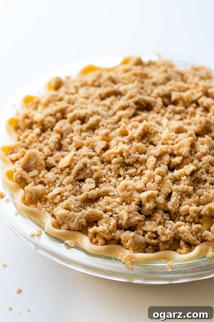 Apple Crumble Pie is made with a tender crust that is filled with juicy, spiced apples and topped with a delicious, buttery streusel topping. This homemade dutch apple pie is easier to make than a classic double-crusted pie and makes the perfect fall dessert! You can even make it a day ahead and re-crisp it just before serving, perfect for Thanksgiving and other special occasions. #applepie #applecrumblepie #dutchapplepie #homemadeapplepie #pierecipes #fallbaking #applecrumbpie #crumbtopping Close-up of a glass baking dish containing an unbaked apple pie with a generous streusel topping, showcasing the perfectly crimped crust edges.