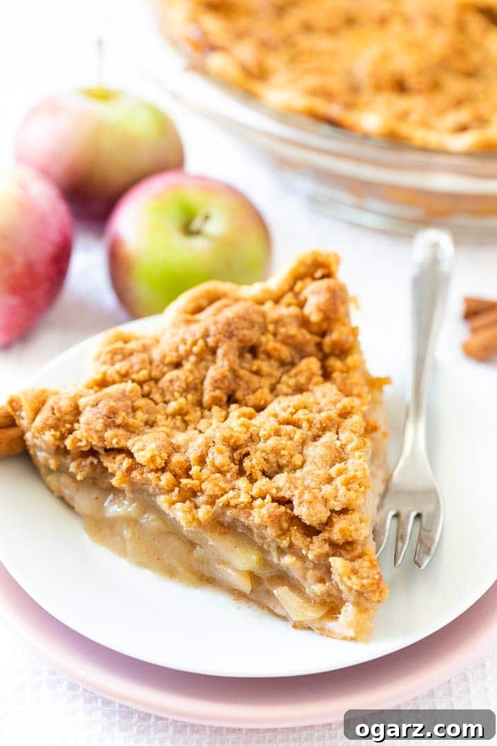 Apple Crumble Pie is made with a tender crust that is filled with juicy, spiced apples and topped with a delicious, buttery streusel topping. This homemade dutch apple pie is easier to make than a classic double-crusted pie and makes the perfect fall dessert! You can even make it a day ahead and re-crisp it just before serving, perfect for Thanksgiving and other special occasions. #applepie #applecrumblepie #dutchapplepie #homemadeapplepie #pierecipes #fallbaking #applecrumbpie #crumbtopping A delectable piece of apple pie with streusel topping, elegantly presented on a white plate atop a pink plate, accompanied by a fork and crowned with a scoop of vanilla ice cream. The scene is artfully garnished with cinnamon sticks and apple slices, with a glass baking dish in the soft background.