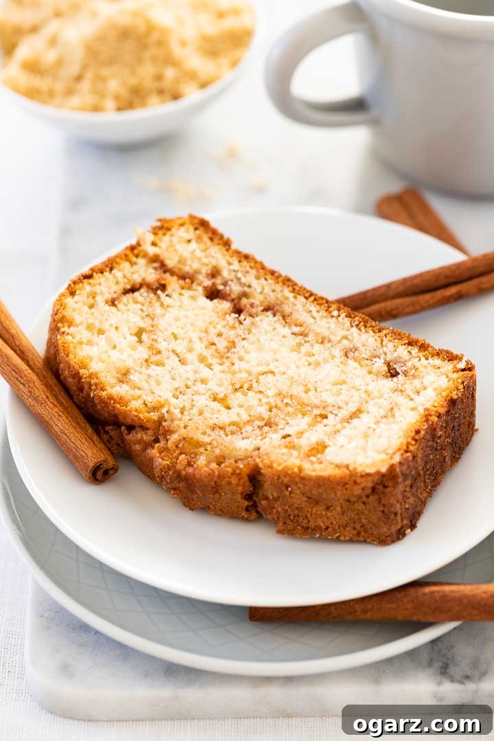 Velvet Cinnamon Swirl 2 A white plate with a slice of cinnamon swirl bread, garnished with some cinnamon sticks and a grey canteen cup and a white bowl with cinnamon sugar mixture in the background.