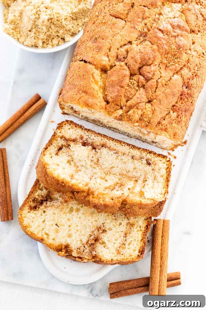 Cinnamon Swirl Bread is so easy to make without yeast and has a delicious cinnamon sugar swirl inside and a crunchy cinnamon sugar crust on top. This moist homemade Cinnamon Bread tastes delicious for breakfast but also makes a great afternoon snack! A quick bread that's perfect for the holidays and keeps fresh for days. #cinnamon #cinnamonswirlbread #cinnamonbread #quickbread #holidaybaking #recipe Top-down view of cinnamon swirl bread on a white rectangular serving platter, with 2 slices cut off and lying down garnished with cinnamon sticks.