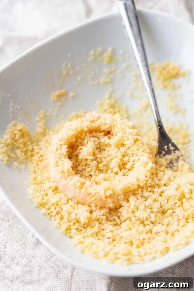 An onion ring being coated in panko breadcrumbs on a white plate with a fork.