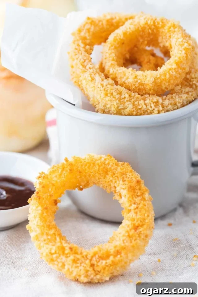 Fried onion rings in a grey canteen cup with parchment paper. There's a small white porcelain bowl with barbecue sauce next to it, a whole raw onion in the background and a single onion ring leaning against it.