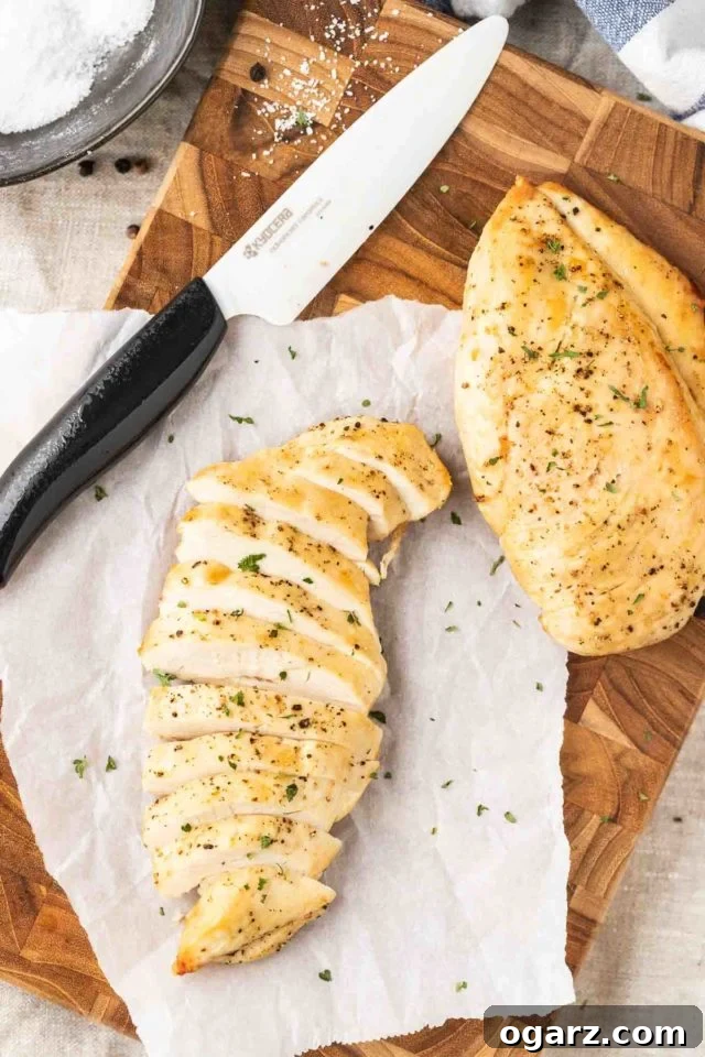Two perfectly cooked, juicy air fryer chicken breasts on a dark wood cutting board with parchment paper, one sliced to reveal its tender interior, alongside a ceramic knife.