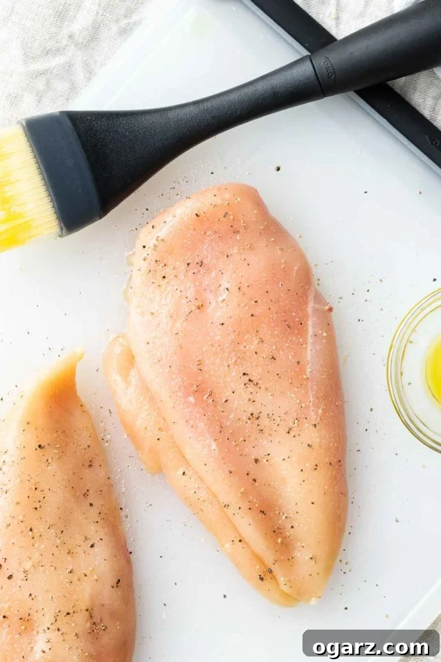 Juicy Air Fryer Chicken Breast rubbed with a simple seasoning and air fried to perfection. This easy and quick chicken breast recipe is perfect for meal prep, salads, and sandwiches! #AirFryerRecipes #BakedChickenBreast Two raw chicken breasts seasoned with salt and pepper on a plastic cutting board, ready for the air fryer, with olive oil and a brush nearby.