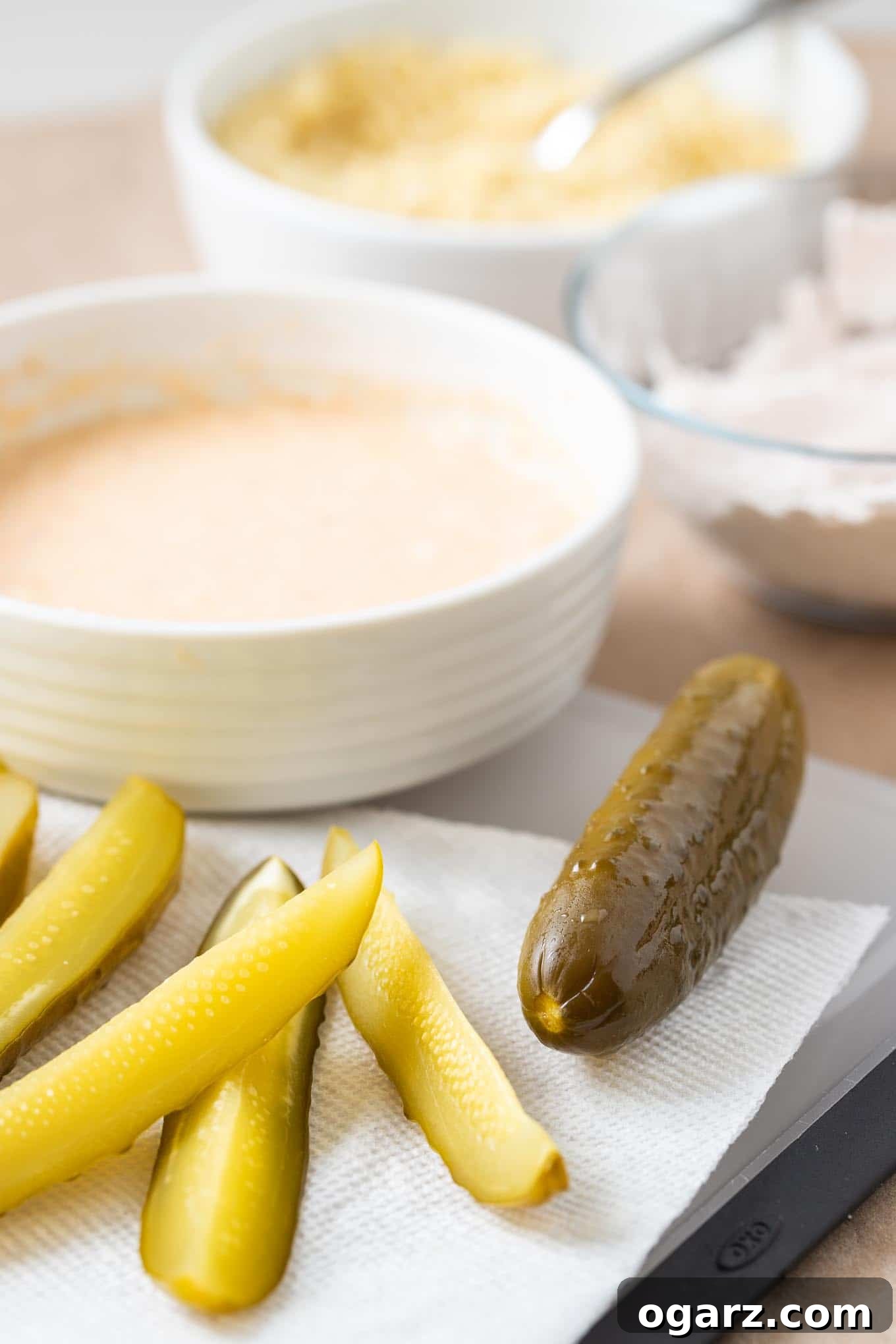 Air Fryer Pickles are super crispy and make a great game day or party snack! Fried Pickles made in the Air Fryer are healthier because they are made with a lot less oil and have a crunchy panko breadcrumbs coating that makes them taste even better than the deep fried original. #AirFryerRecipes #FriedPickles Dill pickle wedges, with a whole dill pickle on a cutting board with a kitchen towel. There\'s a white bowl with batter and a glass bowl with flour in the background.