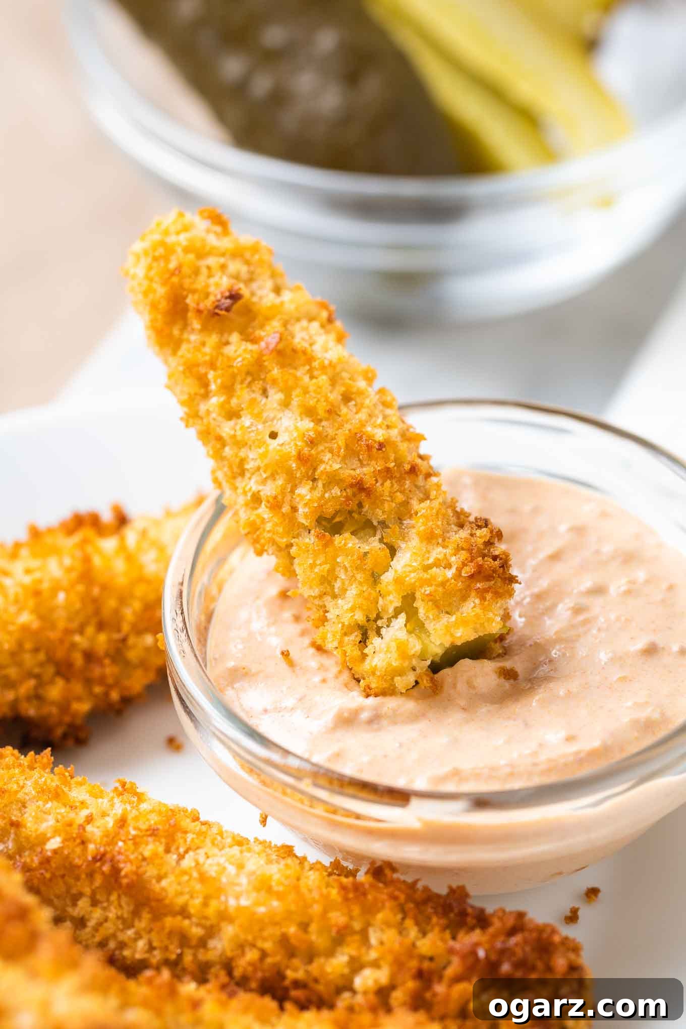Air Fryer Pickles are super crispy and make a great game day or party snack! Fried Pickles made in the Air Fryer are healthier because they are made with a lot less oil and have a crunchy panko breadcrumbs coating that makes them taste even better than the deep fried original. #AirFryerRecipes #FriedPickles A fried dill pickle is dipped into sauce in a small glass bowl, with more fried pickles next to it on a white plate.
