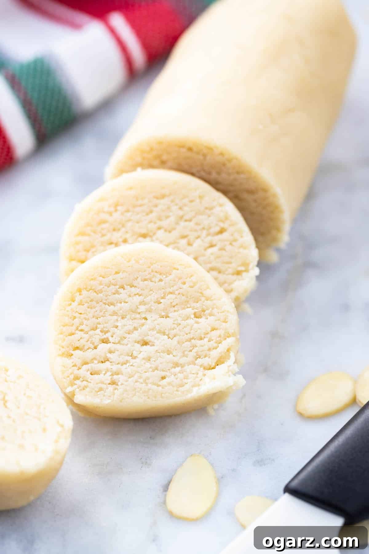 Homemade Marzipan: Superior Flavor, Less Cost. Perfect for Christmas & Easter! Traditional Recipe (No Egg/Corn Syrup). Easy to Make, Flavor, & Dye for Cakes, Cookies, & Candy. A perfectly shaped log of homemade marzipan, with two thin slices cut and leaning against it, set on a elegant grey marble surface. Scattered around are a few whole almonds, hinting at the simple, natural ingredients within this delightful confection. The image title suggests that homemade marzipan offers superior flavor and is more economical than commercial alternatives, making it ideal for various festive decorations, cookies, cakes, figures, and candies. This traditional marzipan recipe, free from egg whites or corn syrup, is easily flavored and dyed, and the guide promises to reveal its simple creation process.