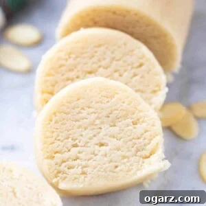 A log of marzipan with two slices cut off and leaning against it on a grey marble surface with some almonds next to it.
