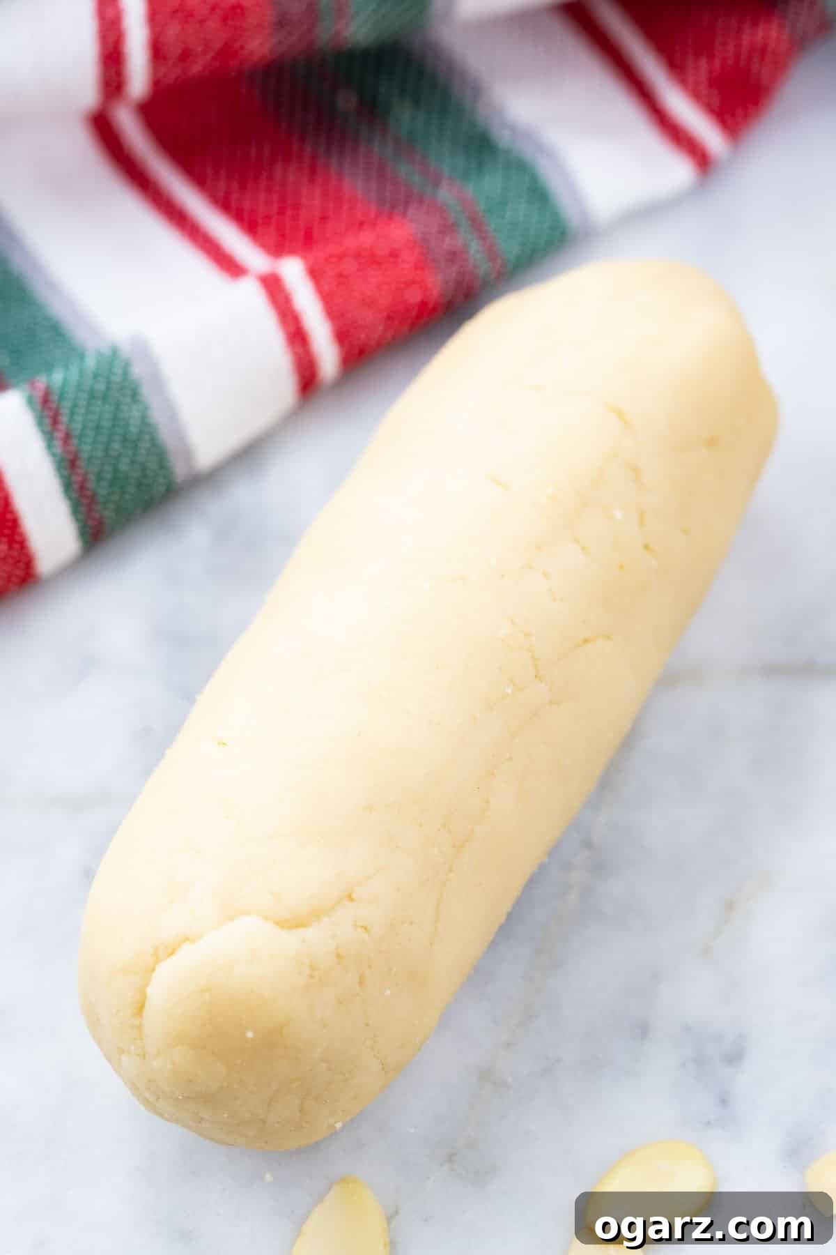 Homemade Marzipan: Superior Flavor, Less Cost. Perfect for Christmas & Easter! Traditional Recipe (No Egg/Corn Syrup). Easy to Make, Flavor, & Dye for Cakes, Cookies, & Candy. A perfectly formed log of rich marzipan rests on a cool grey marble surface, with a vibrant red, white, and green striped dish towel neatly folded in the background. The title reinforces that homemade marzipan offers superior flavor and is more economical than commercial alternatives, making it ideal for various festive decorations, cookies, cakes, figures, and candies. This traditional marzipan recipe, free from egg whites or corn syrup, is easily flavored and dyed, and the guide promises to reveal its simple creation process.