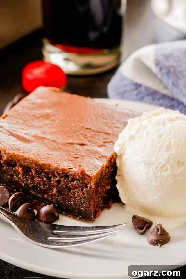 slice of coca-cola cake on a white plate with vanilla ice cream, a fork, and some chocolate chips with a white and blue dish towel and a coca-cola bottle cap in the background