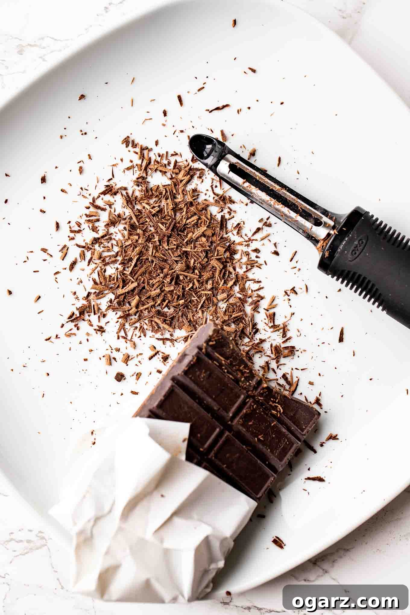Dark Forest Cherry Delight 14 A solid bar of bittersweet baking chocolate and a vegetable peeler on a white plate, prepared for making chocolate shavings for garnish.