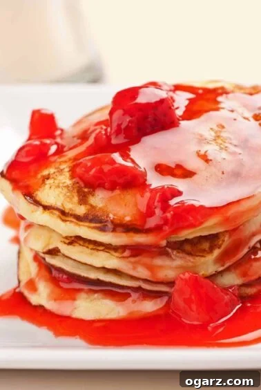 Stack of fluffy ricotta pancakes topped with vibrant strawberry-orange sauce and chunks of fresh strawberries, served on a white plate, ideal for a special brunch.