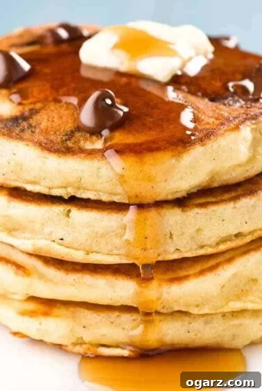 A stack of four chocolate chip pancakes topped with a few extra chocolate chips, a pat of butter, and a drizzle of maple syrup, a delightful sweet-savory brunch option.