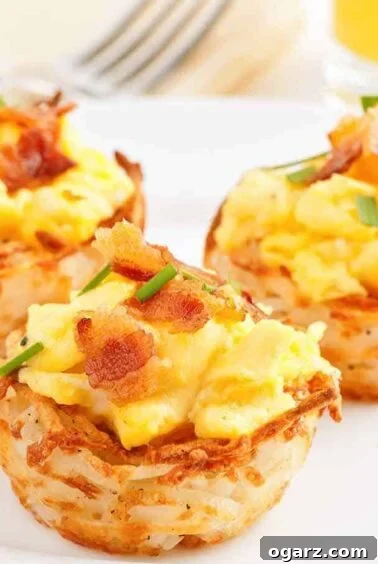 A serving plate with three hash brown potato cups filled with scrambled eggs topped with bacon and garnished with chives, a charming savory brunch item.
