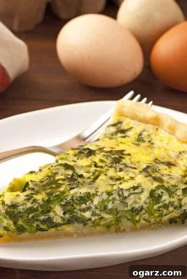 A slice of spinach and cheese quiche on a plate with a fork, fresh eggs and and a tea towel in the background, highlighting a classic and versatile brunch dish.